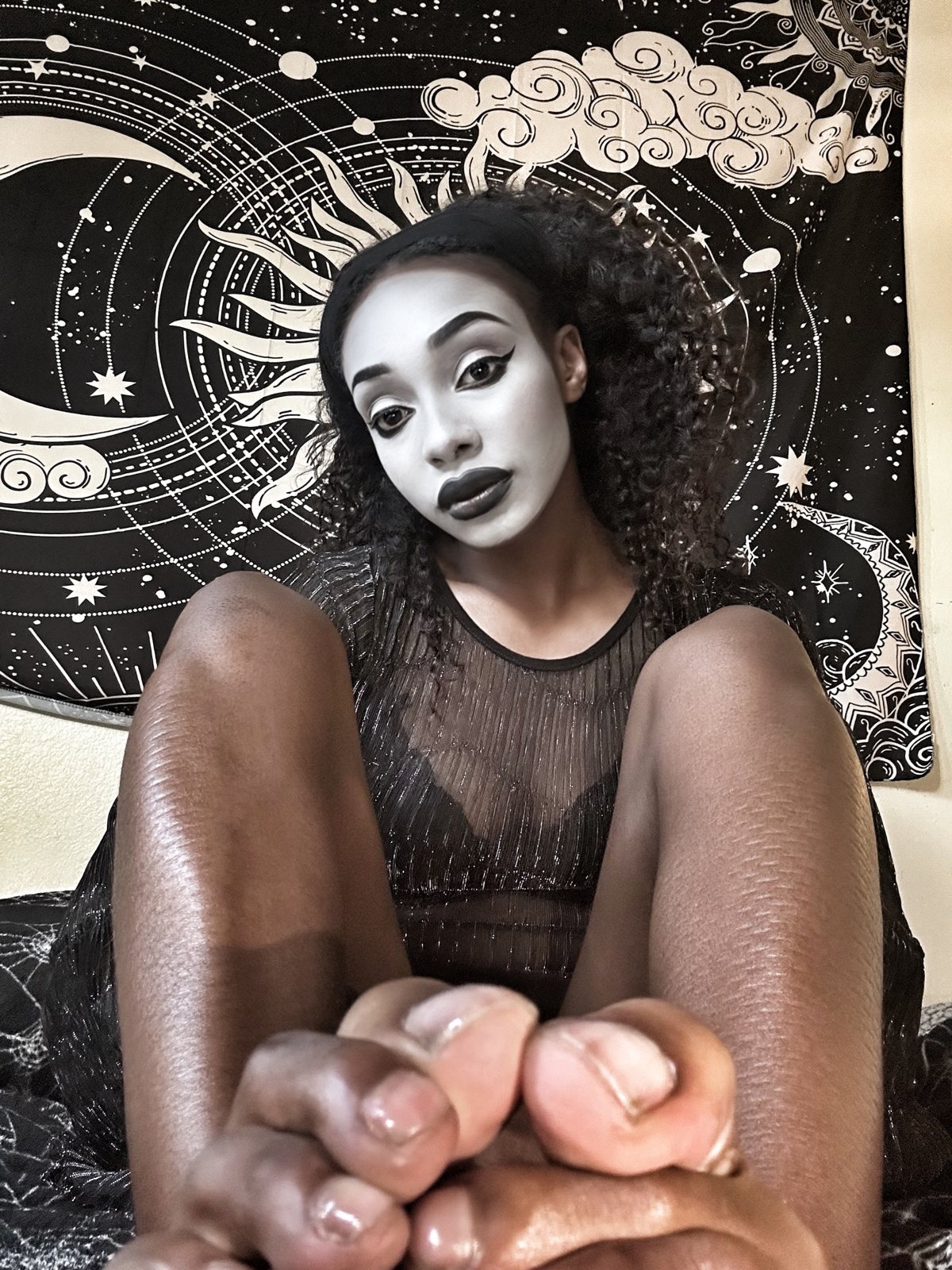 Queen Kenna in white face paint and black lips, bare feet toward the camera, under a sun and moon tapestry